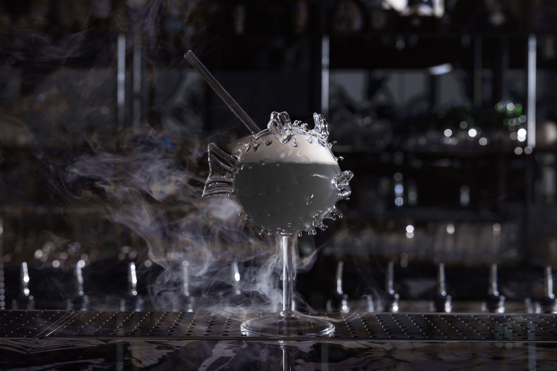 A glass pufferfish cocktail wrapped in dry-ice smoke on the dark bar