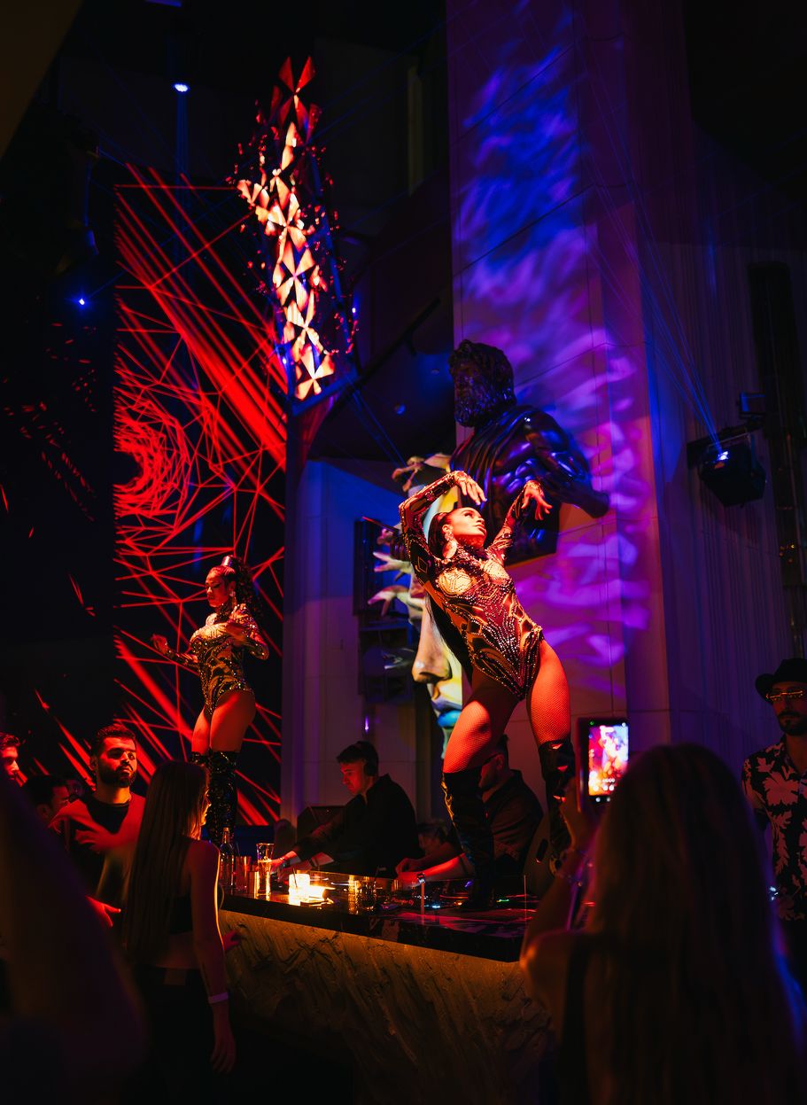 Two dancers in gold on the DJ booth beneath the bronze Zeus, red laser mesh behind